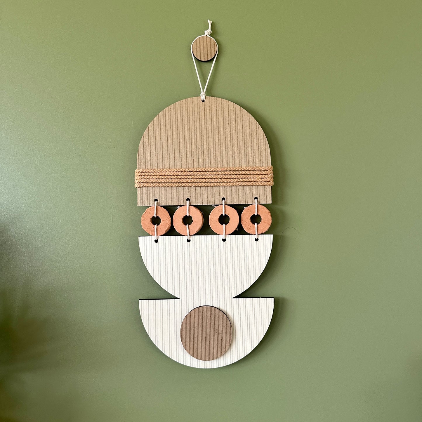 A bohemian style geometric wall hanging made from plywood part painted in beige and part left wood. Cut in semi circles and circles. With terracotta ceramic discs and brown yarn to add texture to the piece. 40cmL x 16cmW