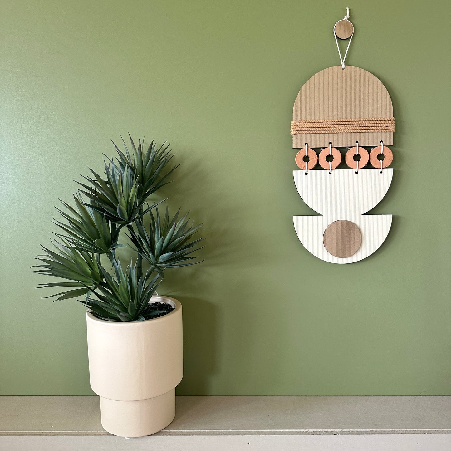 A bohemian style geometric wall hanging made from plywood part painted in beige and part left wood. Cut in semi circles and circles. With terracotta ceramic discs and brown yarn to add texture to the piece. 40cmL x 16cmW