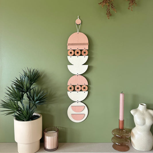 A bohemian style geometric wall hanging made from plywood part painted in pink and part left wood. Cut in semi circles and circles. With terracotta ceramic discs and pink yarn to add texture to the piece. 60cmL x 12cmW