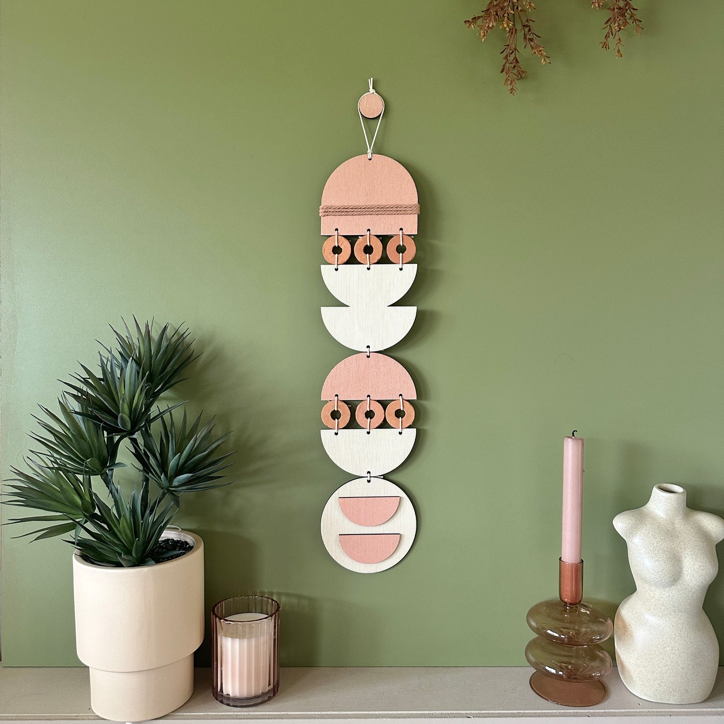 A bohemian style geometric wall hanging made from plywood part painted in pink and part left wood. Cut in semi circles and circles. With terracotta ceramic discs and pink yarn to add texture to the piece. 60cmL x 12cmW