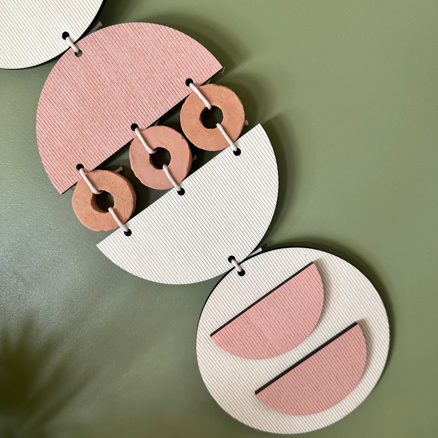 A bohemian style geometric wall hanging made from plywood part painted in pink and part left wood. Cut in semi circles and circles. With terracotta ceramic discs and pink yarn to add texture to the piece. 60cmL x 12cmW