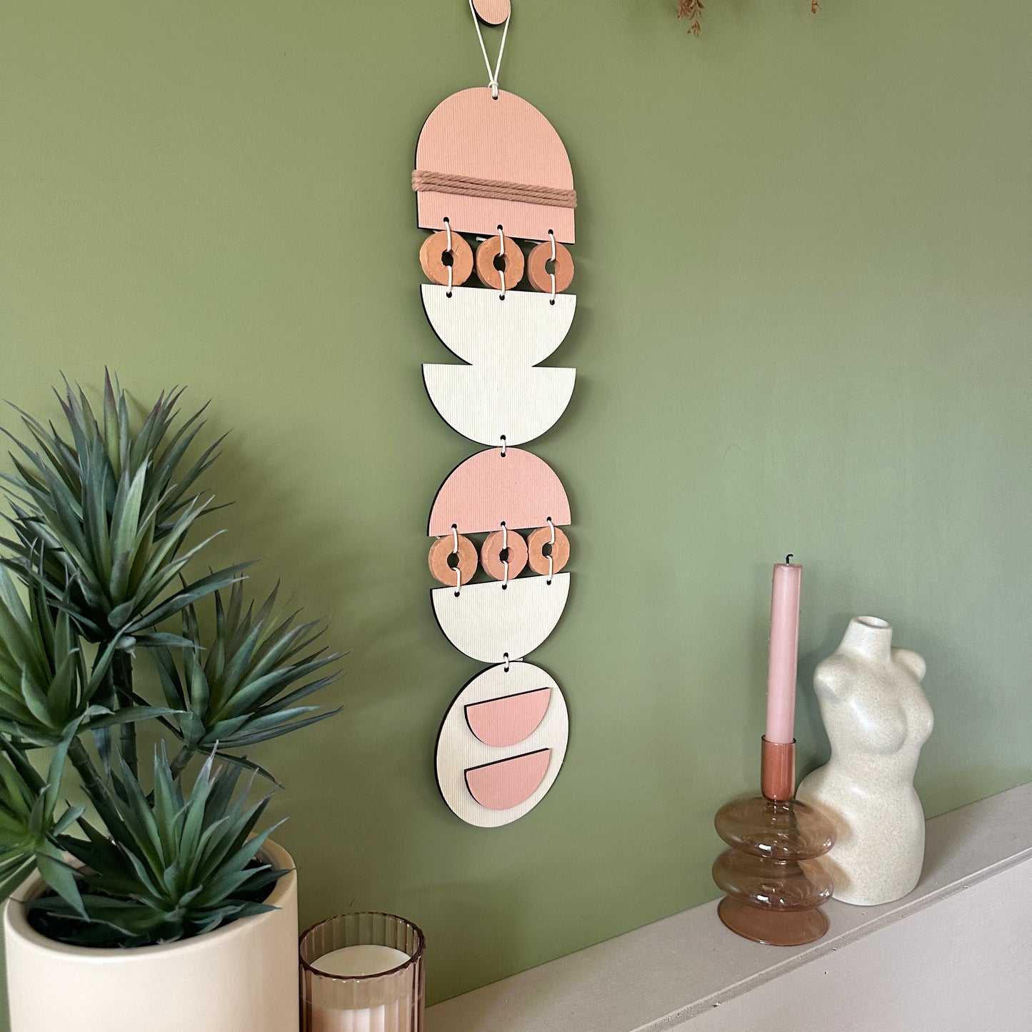 A bohemian style geometric wall hanging made from plywood part painted in pink and part left wood. Cut in semi circles and circles. With terracotta ceramic discs and pink yarn to add texture to the piece. 60cmL x 12cmW