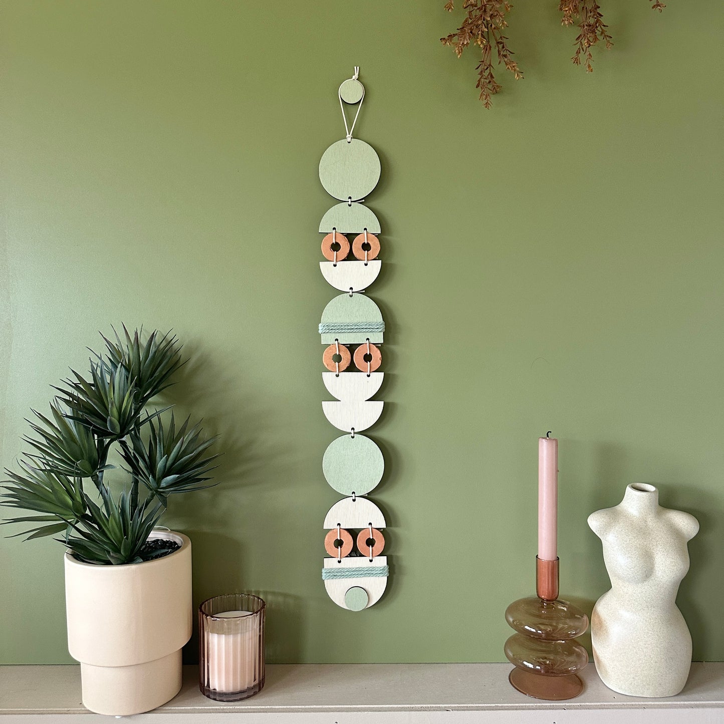 A bohemian style geometric wall hanging made from plywood part painted in green and part left wood. Cut in semi circles and circles. With terracotta ceramic discs and green yarn to add texture to the piece. 68cmL x 8cmW