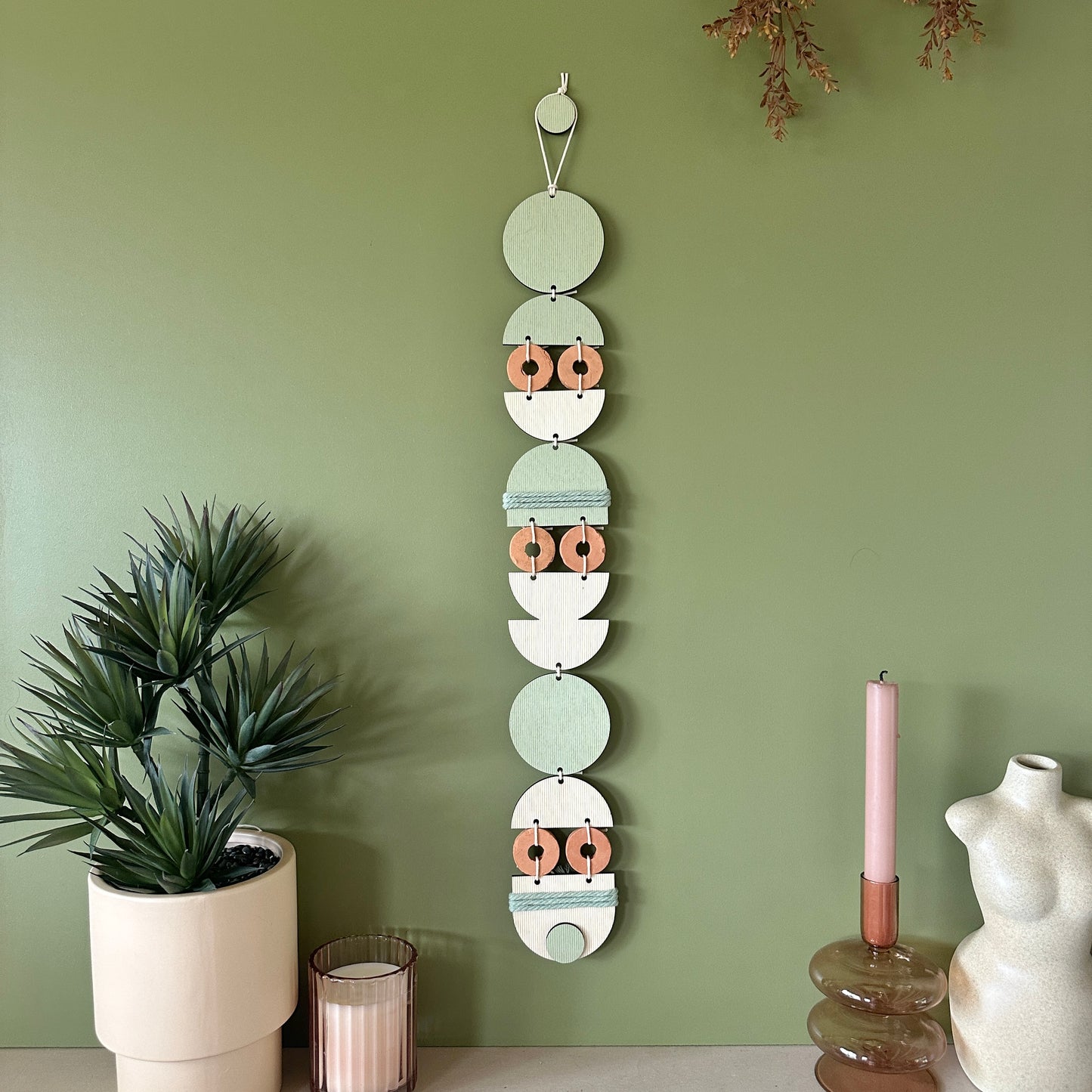 A bohemian style geometric wall hanging made from plywood part painted in green and part left wood. Cut in semi circles and circles. With terracotta ceramic discs and green yarn to add texture to the piece. 68cmL x 8cmW