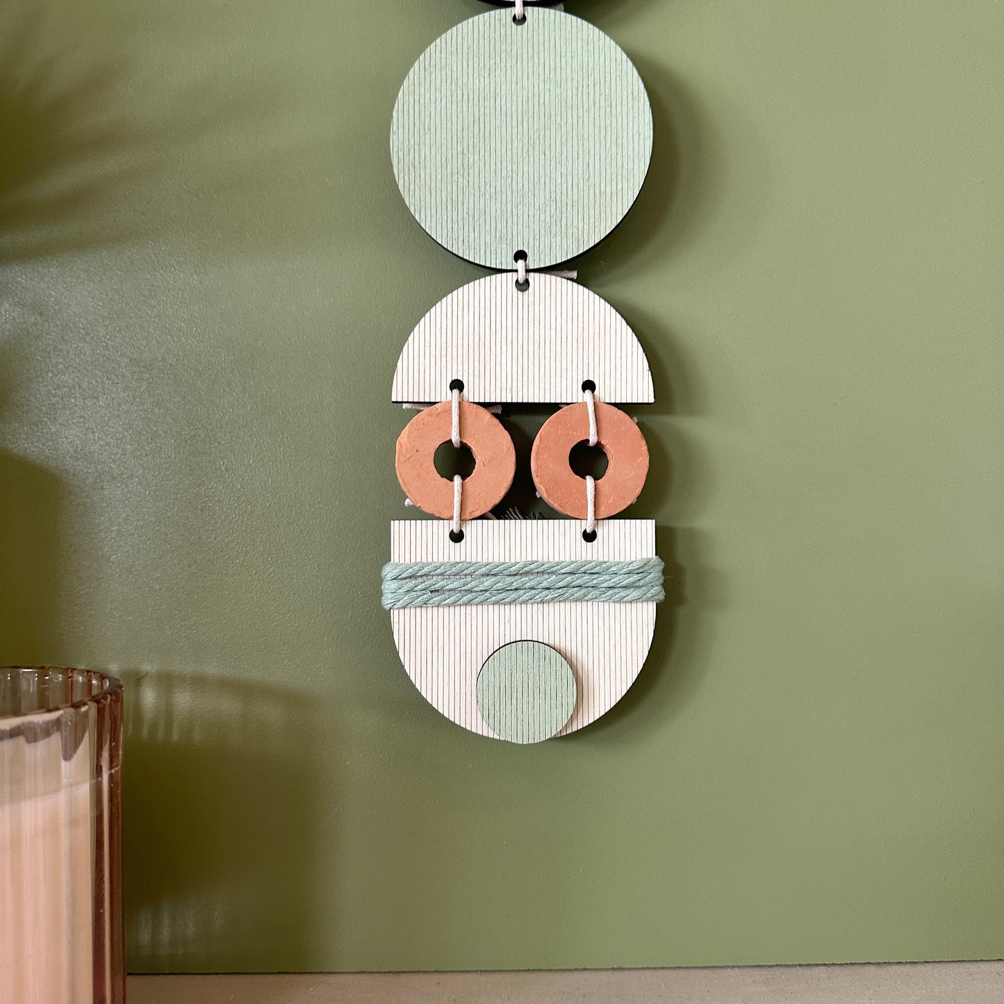 A bohemian style geometric wall hanging made from plywood part painted in green and part left wood. Cut in semi circles and circles. With terracotta ceramic discs and green yarn to add texture to the piece. 68cmL x 8cmW