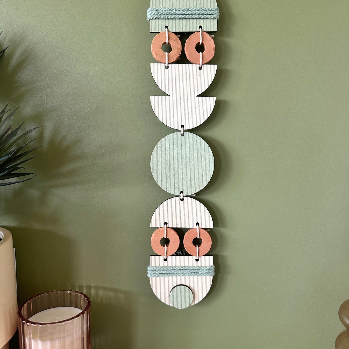 A bohemian style geometric wall hanging made from plywood part painted in green and part left wood. Cut in semi circles and circles. With terracotta ceramic discs and green yarn to add texture to the piece. 68cmL x 8cmW