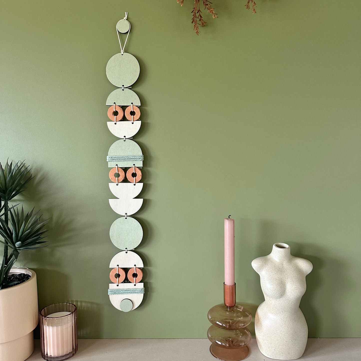 A bohemian style geometric wall hanging made from plywood part painted in green and part left wood. Cut in semi circles and circles. With terracotta ceramic discs and green yarn to add texture to the piece. 68cmL x 8cmW