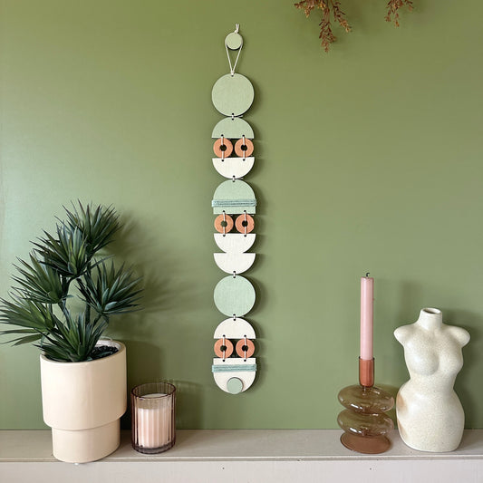 A bohemian style geometric wall hanging made from plywood part painted in green and part left wood. Cut in semi circles and circles. With terracotta ceramic discs and green yarn to add texture to the piece. 68cmL x 8cmW