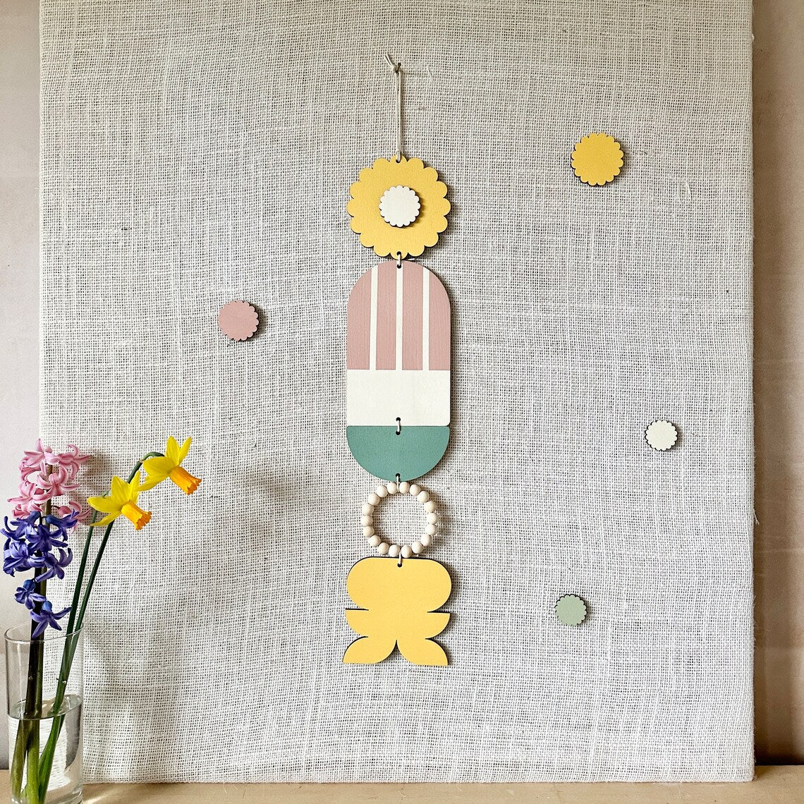 A floral colourful wall hanging is shown in pastel tones. Made from sustainable plywood pieces tied together to create a larger piece. The range includes pieces with green, yellow, blue, pink and brown colours. Some of the wood pieces are flowers.