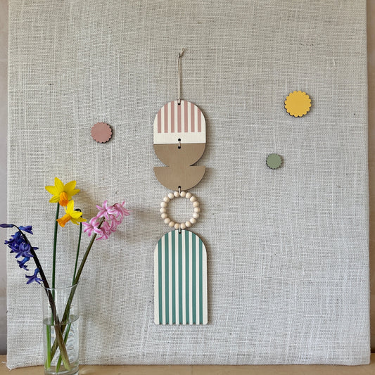 A floral colourful wall hanging is shown in pastel tones. Made from sustainable plywood pieces tied together to create larger piece. The range includes pieces with green, yellow, blue, pink brown colours. Some of the wood pieces are flowers.