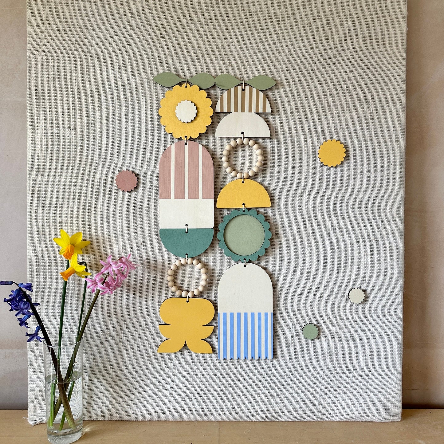 A floral colourful wall hanging is shown in pastel tones. Made from sustainable plywood pieces tied together to create a larger piece. The range includes pieces with green, yellow, blue, pink and brown colours. Some of the wood pieces are flowers.