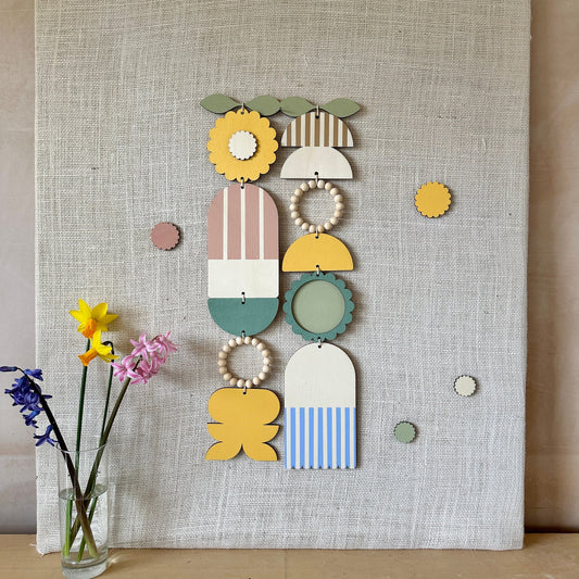 A floral colourful wall hanging is shown in pastel tones. Made from sustainable plywood pieces tied together to create a larger piece. The range includes pieces with green, yellow, blue, pink and brown colours. Some of the wood pieces are flowers.