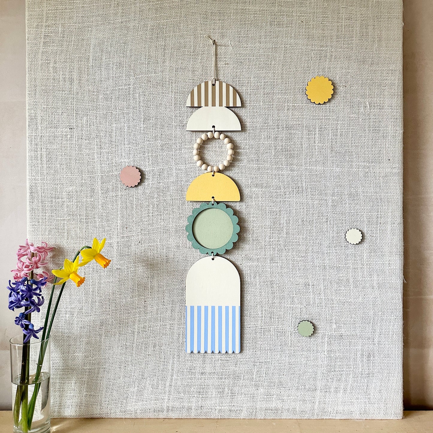 A floral colourful wall hanging is shown in pastel tones. Made from sustainable plywood pieces tied together to create a larger piece. The range includes pieces with green, yellow, blue, pink and brown colours. Some of the wood pieces are flowers.