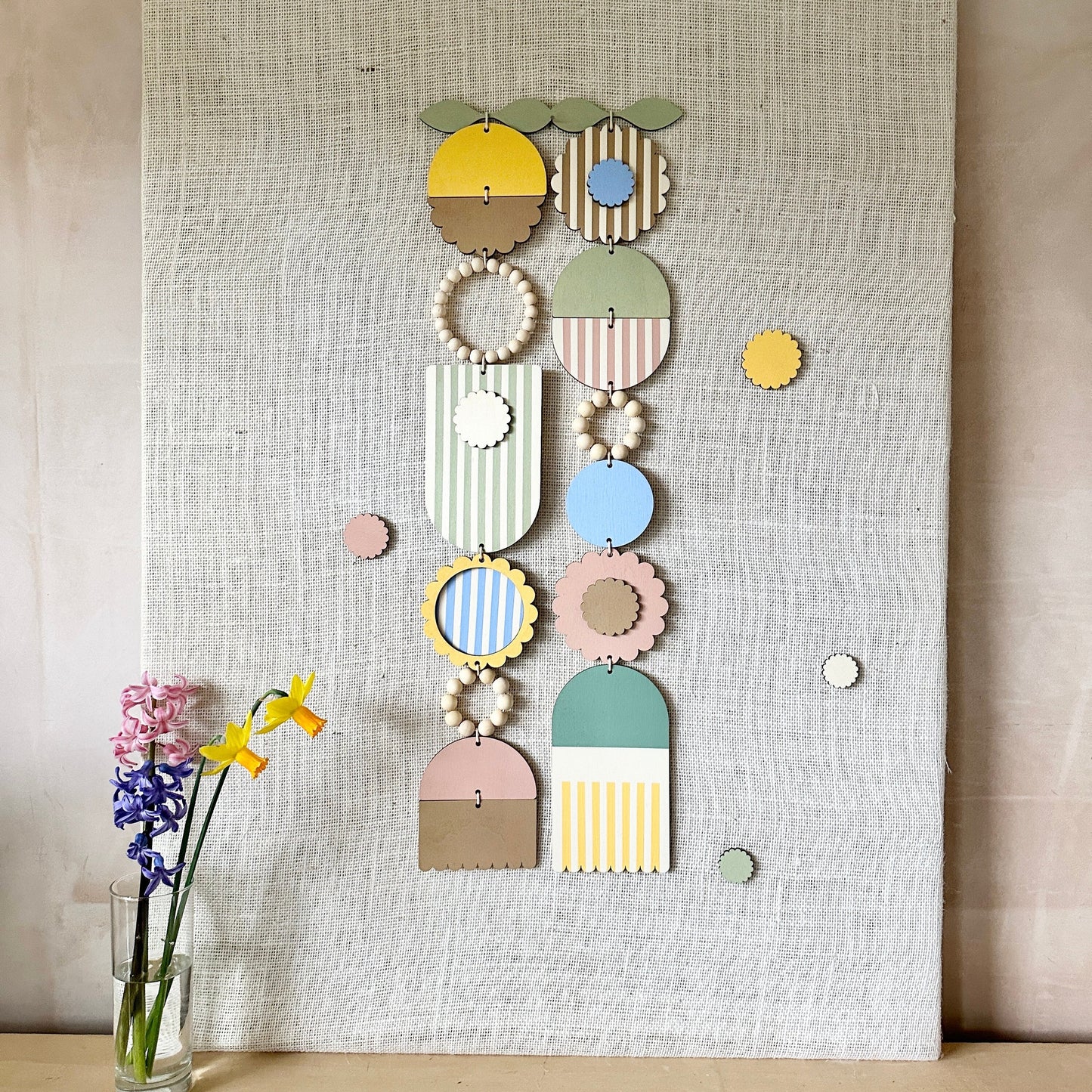 A floral colourful wall hanging is shown in pastel tones. Made from sustainable plywood pieces tied together to create a larger piece. The range includes pieces with green, yellow, blue, pink and brown colours. Some of the wood pieces are flowers.