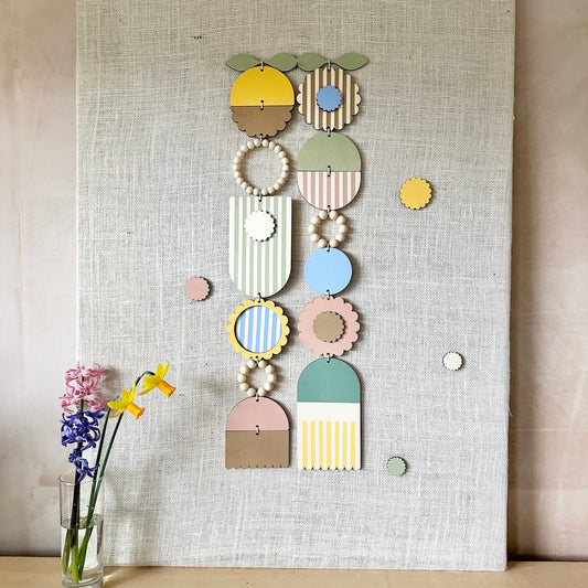 A floral colourful wall hanging is shown in pastel tones. Made from sustainable plywood pieces tied together to create a larger piece. The range includes pieces with green, yellow, blue, pink and brown colours. Some of the wood pieces are flowers.