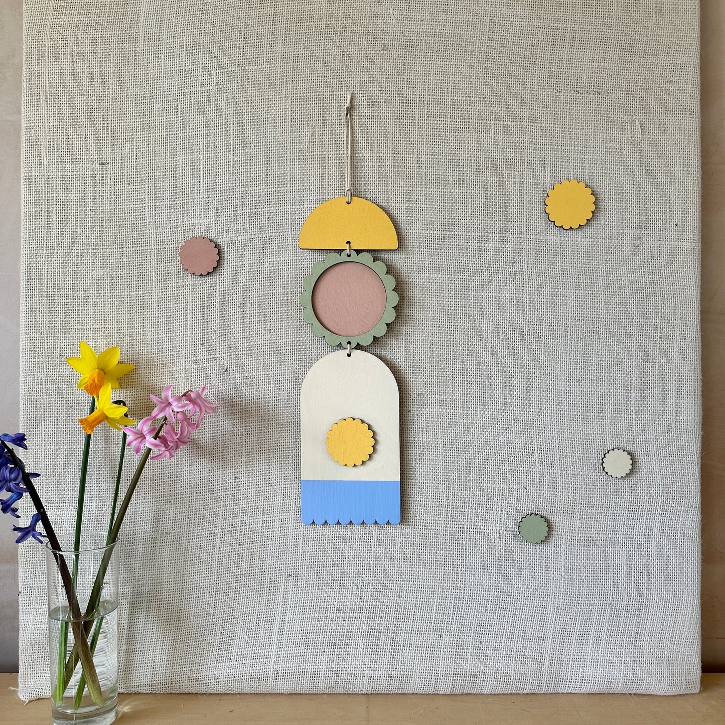 A floral colourful wall hanging is shown in pastel tones. Made from sustainable plywood pieces tied together to create larger piece. The range includes pieces with green, yellow, blue, pink brown colours. Some of the wood pieces are flowers.