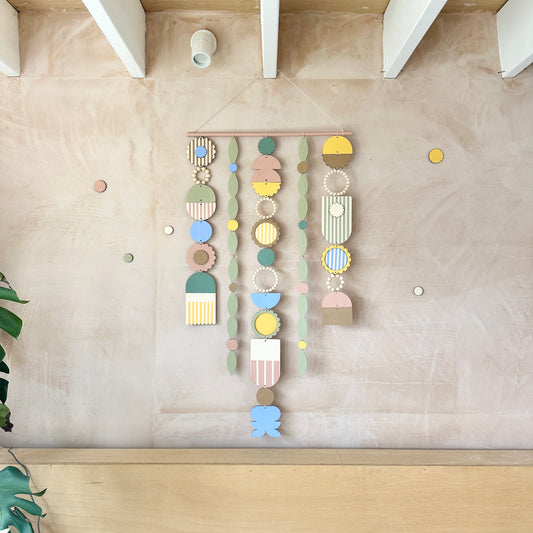 A floral colourful wall hanging is shown in pastel tones. Made from sustainable plywood pieces tied together to create a larger piece. The range includes pieces with green, yellow, blue, pink and brown colours. Some of the wood pieces are flowers.