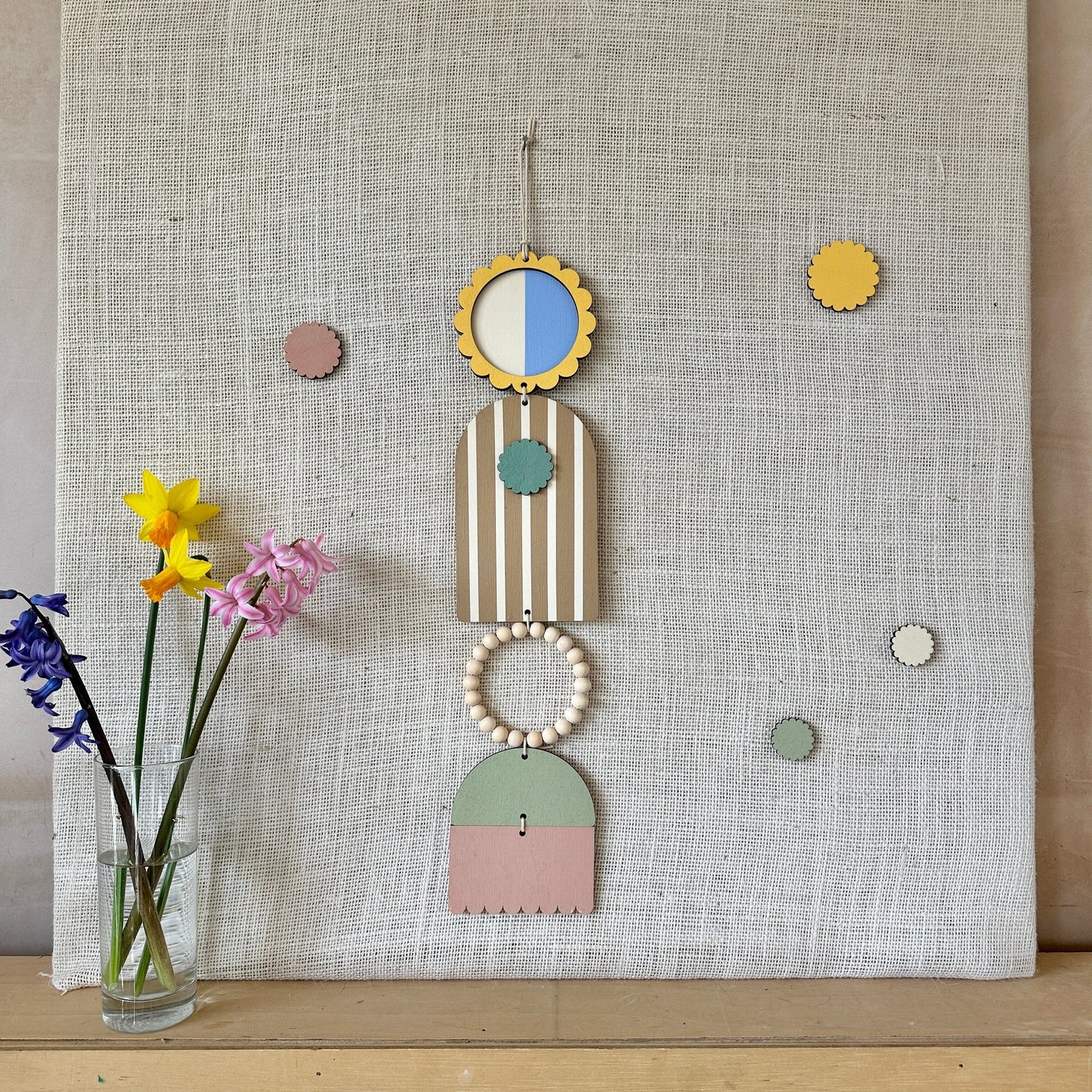 A floral colourful wall hanging is shown in pastel tones. Made from sustainable plywood pieces tied together to create larger piece. The range includes pieces with green, yellow, blue, pink brown colours. Some of the wood pieces are flowers.