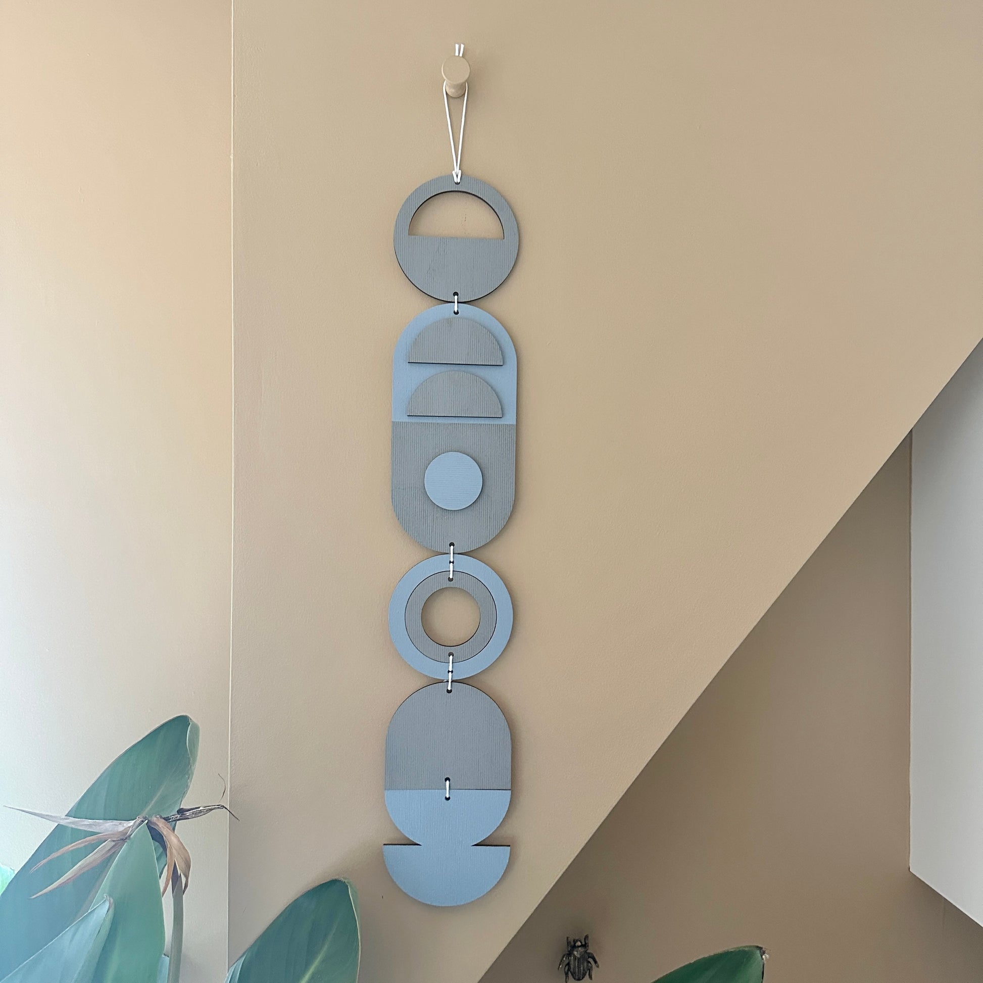 A light blue and grey handmade geometric wood wall hanging. In a modern Bauhaus art style. Featuring a textured effect through layer wood pieces and laser etching. Measuring 68cm in length x 10cm wide