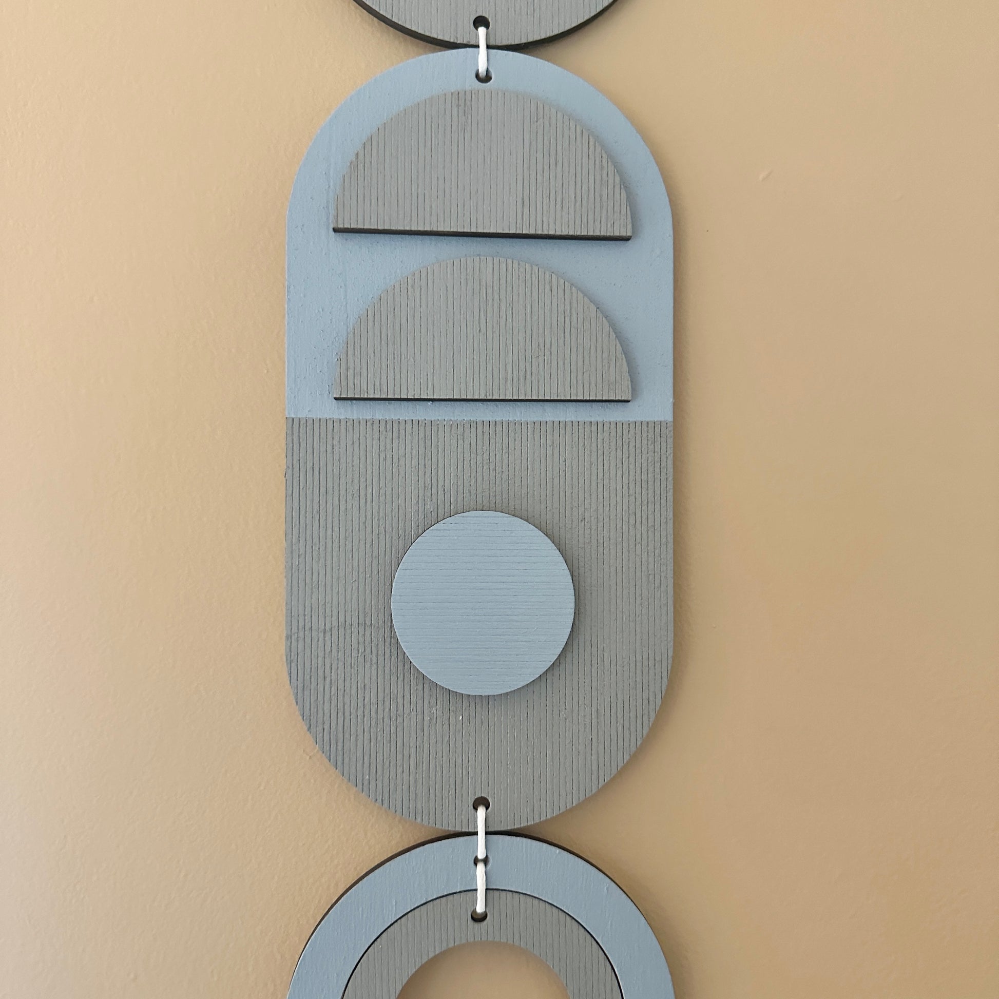 A light blue and grey handmade geometric wood wall hanging. In a modern Bauhaus art style. Featuring a textured effect through layer wood pieces and laser etching. Measuring 68cm in length x 10cm wide