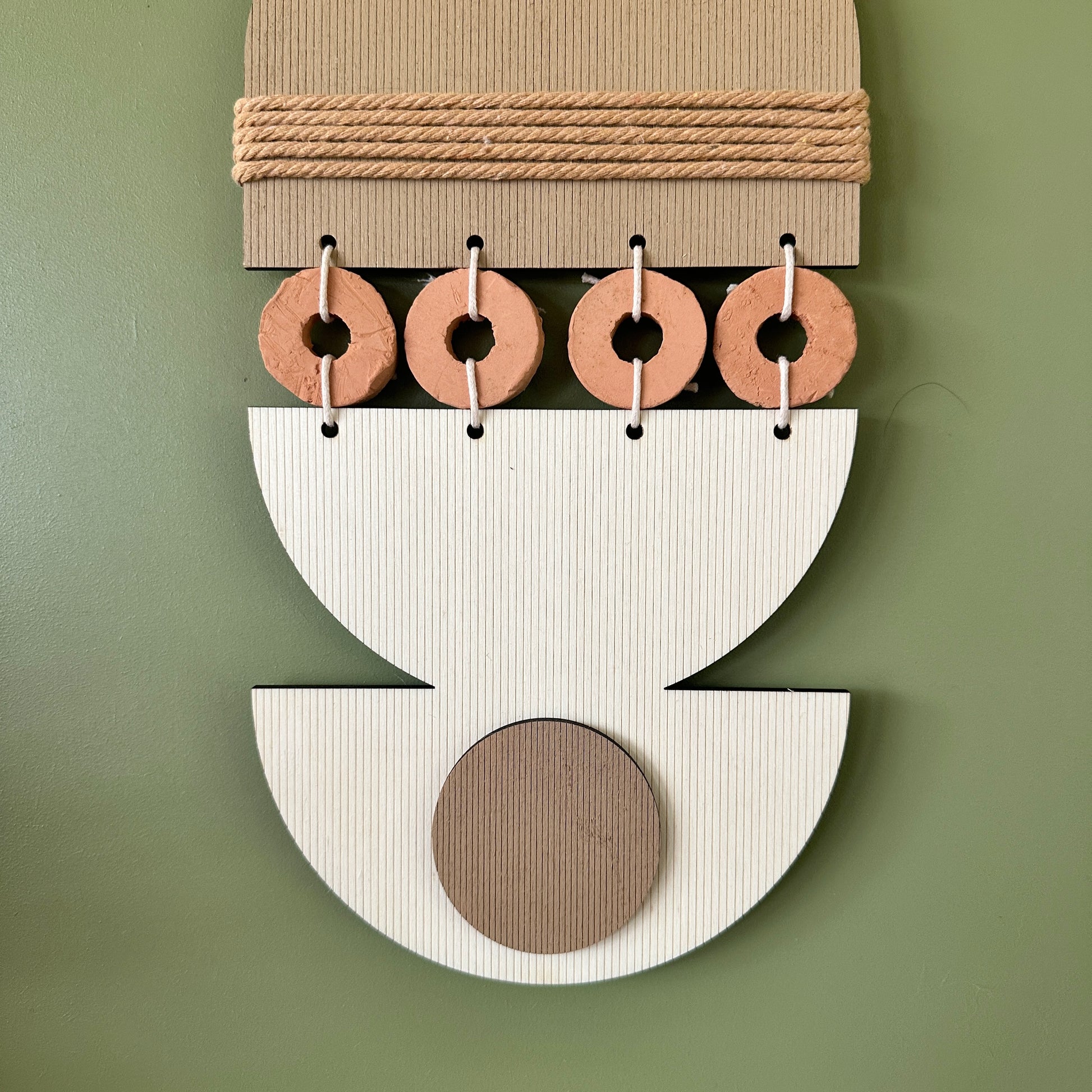 A bohemian style geometric wall hanging made from plywood part painted in beige and part left wood. Cut in semi circles and circles. With terracotta ceramic discs and brown yarn to add texture to the piece. 40cmL x 16cmW