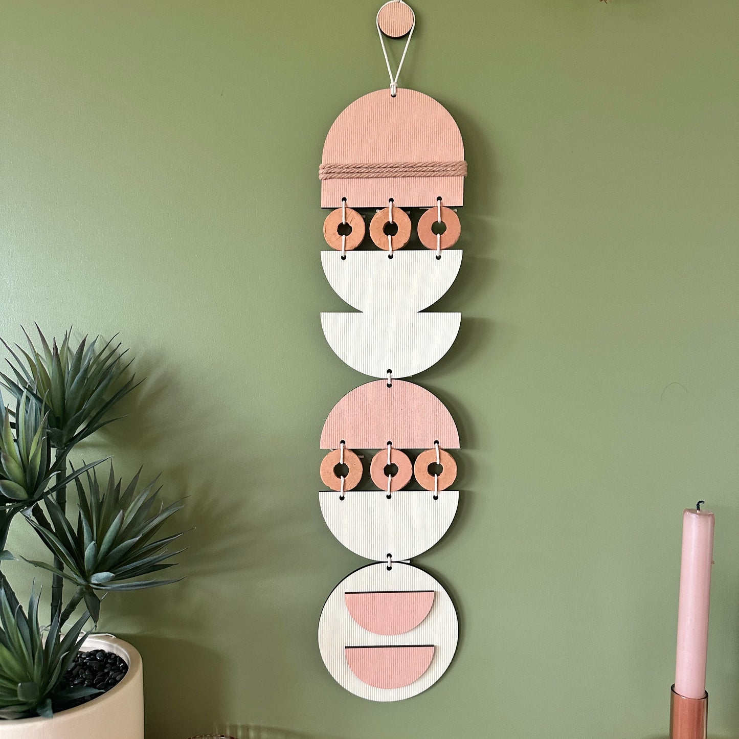 A bohemian style geometric wall hanging made from plywood part painted in pink and part left wood. Cut in semi circles and circles. With terracotta ceramic discs and pink yarn to add texture to the piece. 60cmL x 12cmW