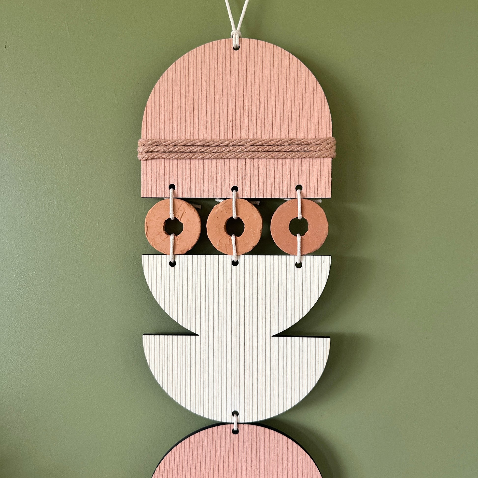 A bohemian style geometric wall hanging made from plywood part painted in pink and part left wood. Cut in semi circles and circles. With terracotta ceramic discs and pink yarn to add texture to the piece. 60cmL x 12cmW