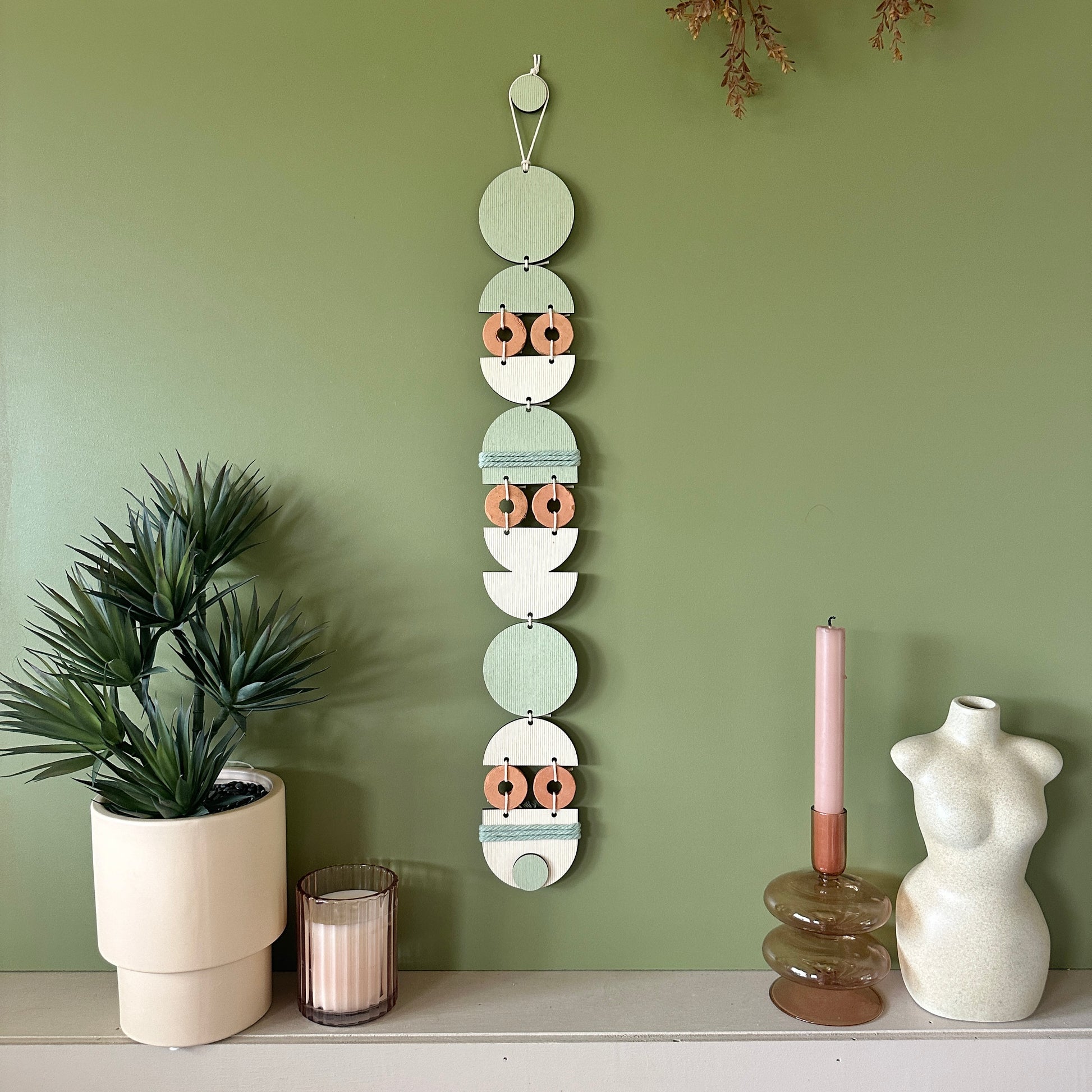 A bohemian style geometric wall hanging made from plywood part painted in green and part left wood. Cut in semi circles and circles. With terracotta ceramic discs and green yarn to add texture to the piece. 68cmL x 8cmW