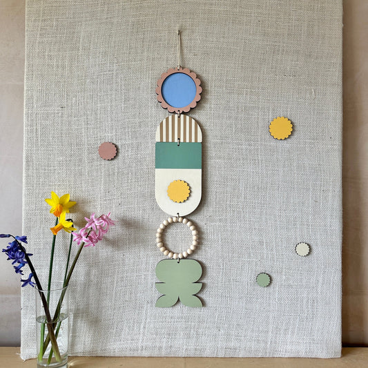 A floral colourful wall hanging is shown in pastel tones. Made from sustainable plywood pieces tied together to create larger piece. The range includes pieces with green, yellow, blue, pink brown colours. Some of the wood pieces are flowers.