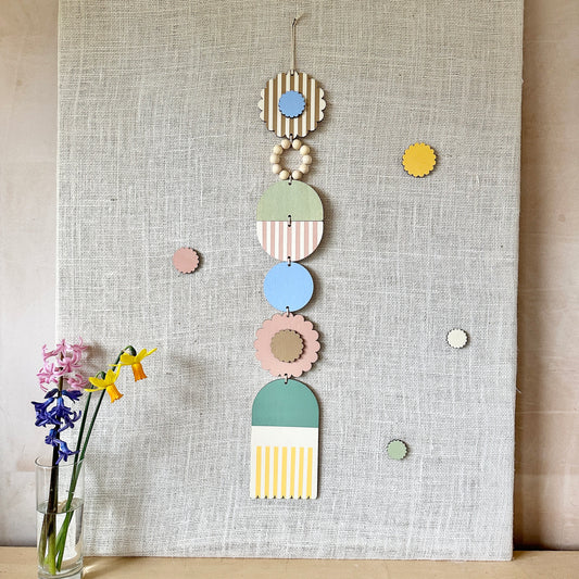 A floral colourful wall hanging is shown in pastel tones. Made from sustainable plywood pieces tied together to create a larger piece. The range includes pieces with green, yellow, blue, pink and brown colours. Some of the wood pieces are flowers.