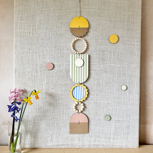 A floral colourful wall hanging is shown in pastel tones. Made from sustainable plywood pieces tied together to create a larger piece. The range includes pieces with green, yellow, blue, pink and brown colours. Some of the wood pieces are flowers.