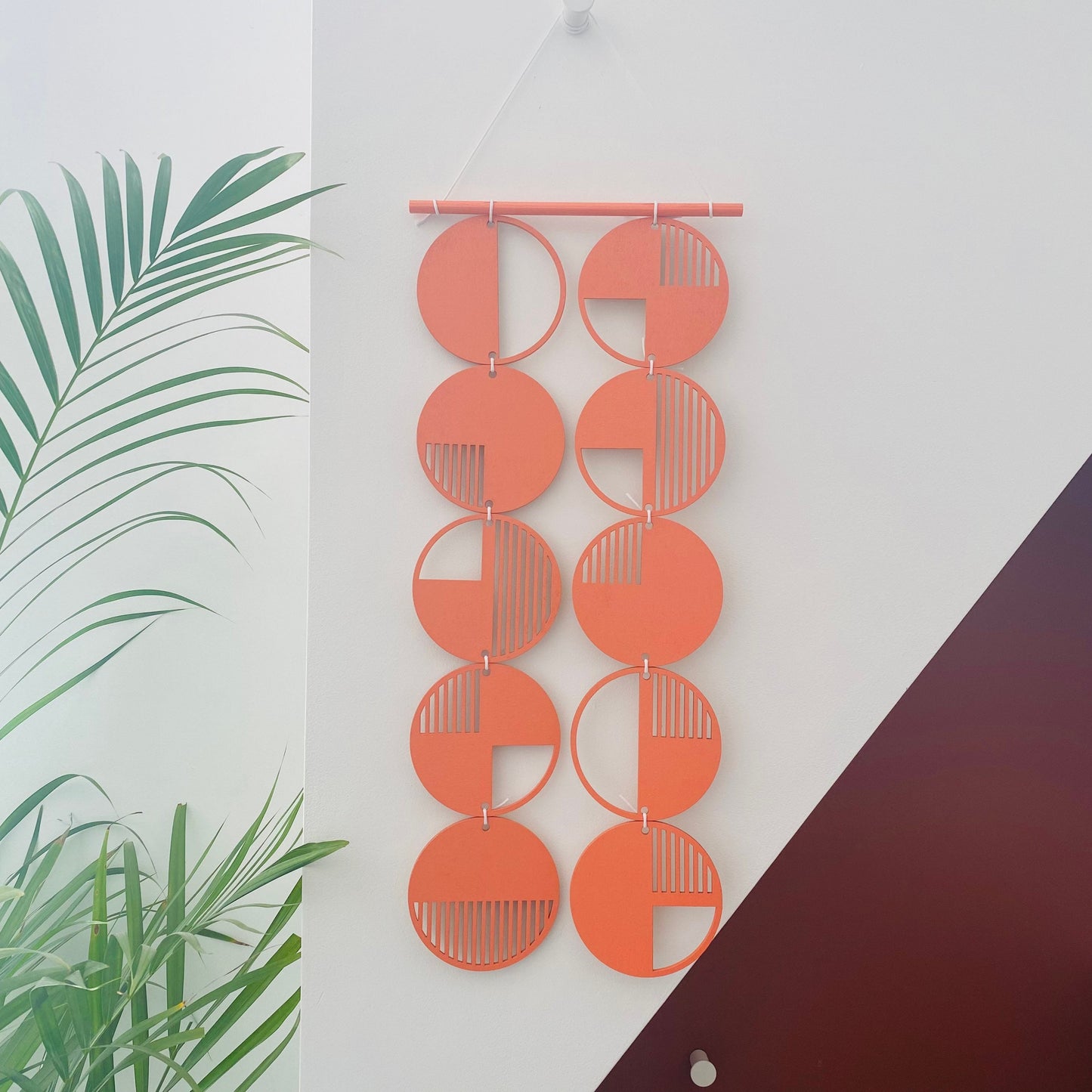 Orange wall art made from 10 wood discs tied to each other 2 rows of 5. With laser cut geometric patterns. 